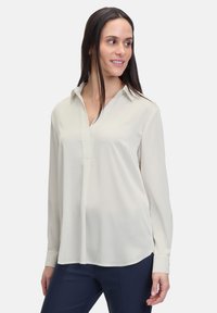 Helle beige Bluse mit langen Ärmeln, aus weichem Stoff, mit Kragen, Knopfverschluss und abgerundetem Saum.