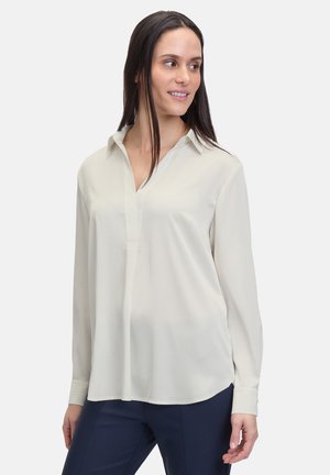 Blusa lunga beige chiaro a maniche lunghe, realizzata in tessuto morbido, con colletto, patta con bottoni e orlo curvo.