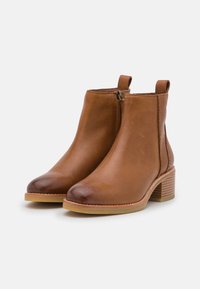 Botas de tobillo de cuero marrón con punta ligeramente redondeada, cremallera lateral y un tacón apilado texturizado. Superficie lisa con un detalle mínimo.