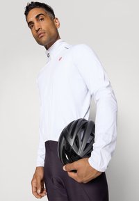 Weißer Radjacke mit hohem Kragen, Reißverschluss und grauen Akzenten; schwarzer Fahrradhelm in der Hand; figurbetonte dunkle Leggings.