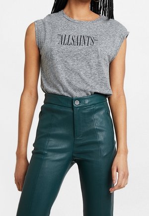 Nainen, jolla on harmaa hihaton AllSaints T-paita, joka on sullottu korkeavyötäröisiin tummanvihreisiin nahkahousuihin, seisoo käsivarret rentoina.