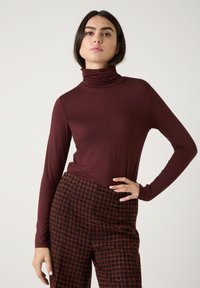 Burgunderfarbener Rollkragenpullover aus glattem, dehnbarem Stoff, kombiniert mit hoch taillierten karierten Hosen in Rot und Schwarz mit einem schachbrettartigen Muster.