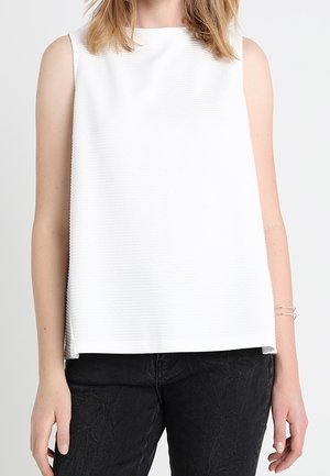 Femme portant un haut blanc sans manches et un pantalon noir texturé, debout devant un fond clair uni.