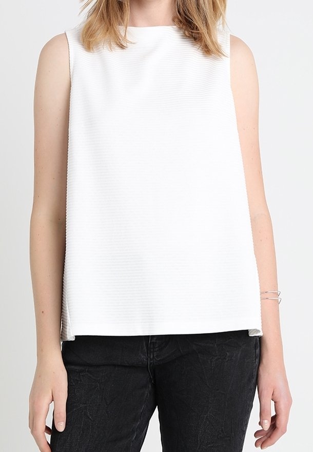Femme portant un haut blanc sans manches et un pantalon noir texturé, debout devant un fond clair uni.