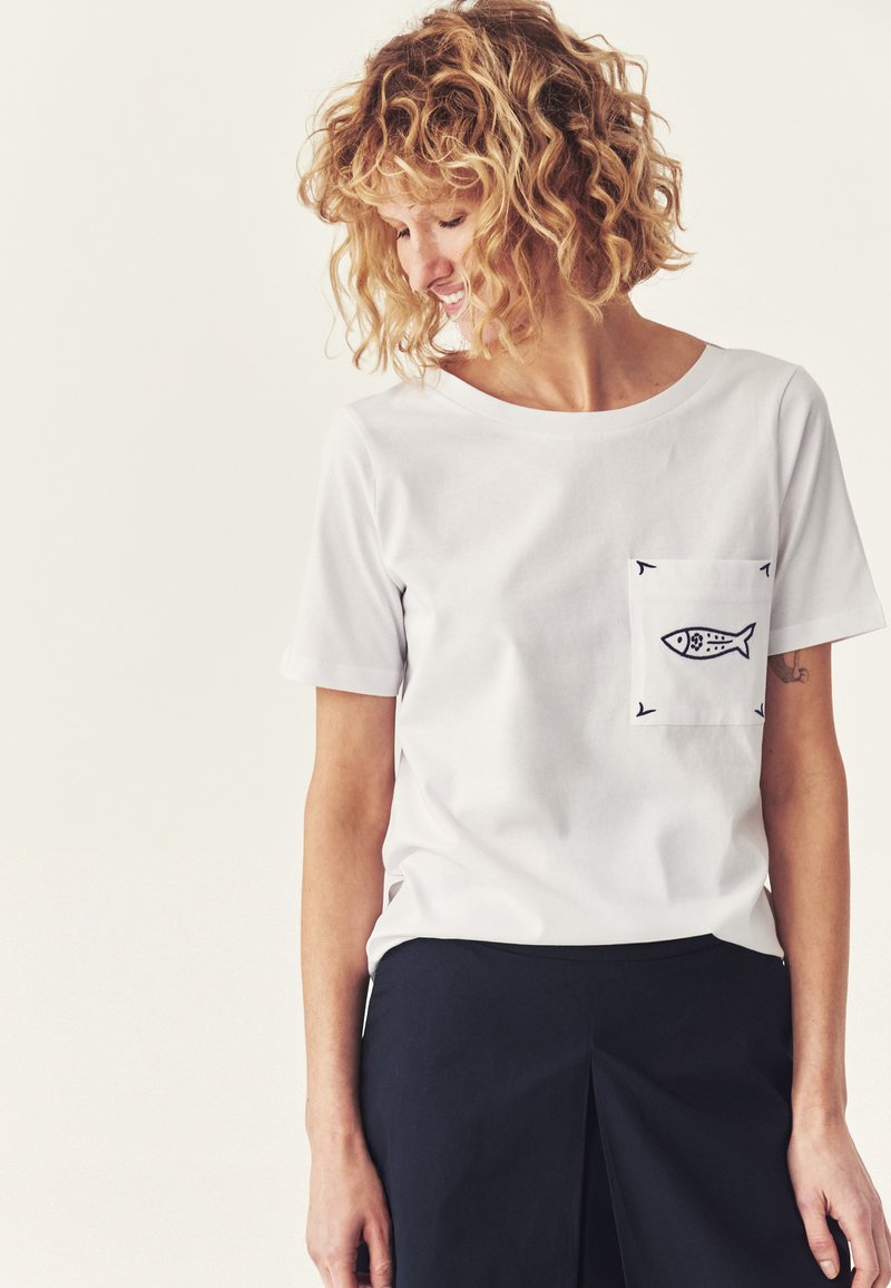 Personne aux cheveux bouclés portant une chemise blanche à manches courtes avec un dessin noir de poisson sur la poche, associée à un pantalon sombre, regardant vers le bas et souriant.