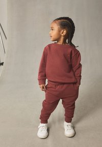 Sweatshirt bordeaux et joggings assortis en tissu doux. Comprend des poignets côtelés et un logo brodé. Associés à des baskets blanches.
