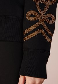 Felpa nera con design ricamato in oro che presenta intricate spirali e svolazzi sulla manica. Tessuto a coste e finitura del polsino.