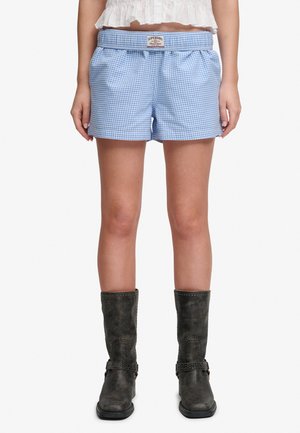 Vrouw draagt blauw-witte geruite shorts met elastische tailleband en zwarte mid-kuitlaarzen, staand tegen een witte achtergrond.