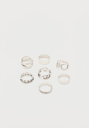 Zeven zilveren ringen met verschillende ontwerpen, waaronder effen banden, kettinkjes en decoratieve patronen, gerangschikt op een witte achtergrond.