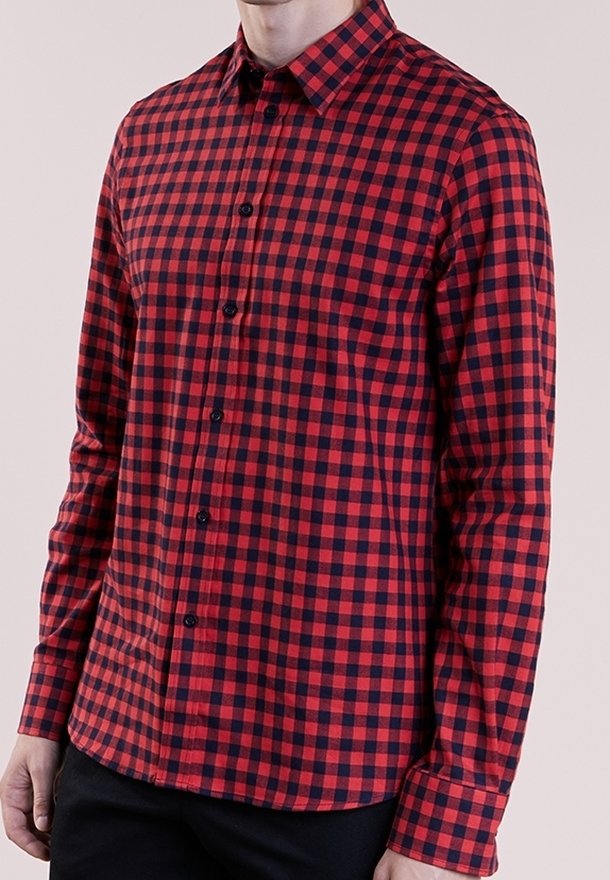 Hombre con una camisa de botones de manga larga ajustada en cuadros rojos y negros, con pantalones negros, de pie contra un fondo sencillo.