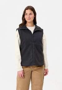 Dunkelgraue Fleeceweste mit einem Kragen, Knopfleiste vorne und strukturierter Stoff. Wird über einem hellen Langarmshirt getragen, kombiniert mit beigen Hosen.