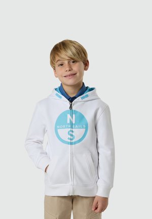 Niño rubio sonriendo, vistiendo una sudadera blanca con cremallera con el logo azul de North Sails y pantalones beige, de pie contra un fondo claro y liso.