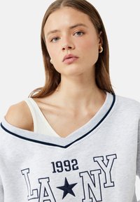 Vaaleanharmaa huppari, jossa on leveä V-pääntie ja tummansininen reunus. Kyljessä on rohkea teksti "LA NY 1992" ja tummansiniset tähti kuvio. Pehmeä rakenne näkyy selvästi.