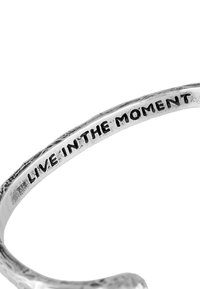 Silver metal cuff bracelet with a textured surface and engraved phrase "LIVE IN THE MOMENT" on the inner side.