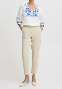 Blusa bianca ricamata con motivi floreali blu, collo a V e maniche a tre quarti, abbinata a pantaloni beige a gamba affusolata e décolleté bianchi a punta.