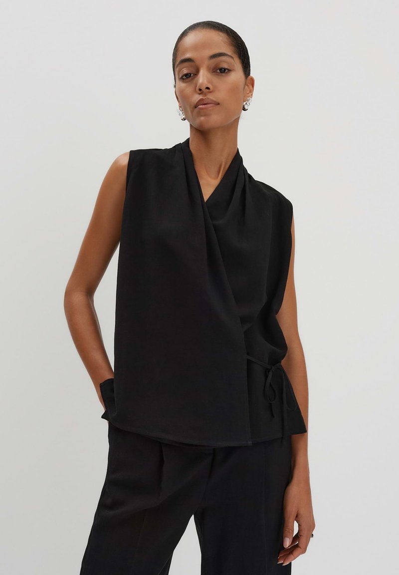 Haut sans manches noir avec un design drapé, fabriqué en tissu doux. Présente une fermeture à lien sur le côté et une coupe décontractée. Associé à un pantalon noir.