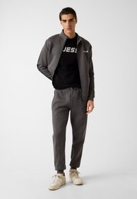 Herren-Sportbekleidungsset mit einer grauen Zip-Up-Jacke und passenden Joggern mit schwarzen Akzenten. Das Ensemble umfasst ein schwarzes T-Shirt mit Logo.