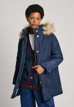 Cappotto imbottito blu navy con cappuccio foderato in pelliccia, dotato di grandi bottoni, cerniera frontale e due tasche laterali. Indossato sopra un maglione di maglia.