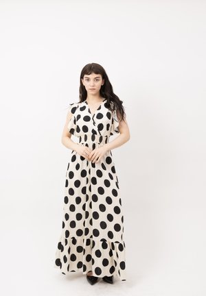 Mujer con cabello largo y oscuro, vistiendo un vestido maxi blanco sin mangas con grandes lunares negros, de pie frente a un fondo blanco liso.