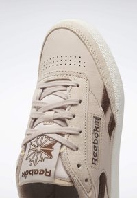 Baskets Reebok beiges avec embout perforé, lacets beiges, panneaux en daim et cuir, et logos de la marque sur la languette et le côté.