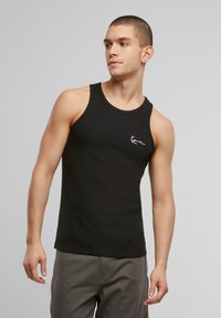 Schwarzes Tanktop aus gerippter Stoff mit Rundhalsausschnitt. Besitzt ein kleines weißes Logo auf der Brust. Das Modell trägt lässige graue Shorts.