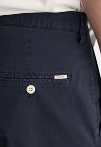 Pantalones azul marino hechos de tela suave con un bolsillo trasero, con un botón blanco y una etiqueta de la marca GANT.
