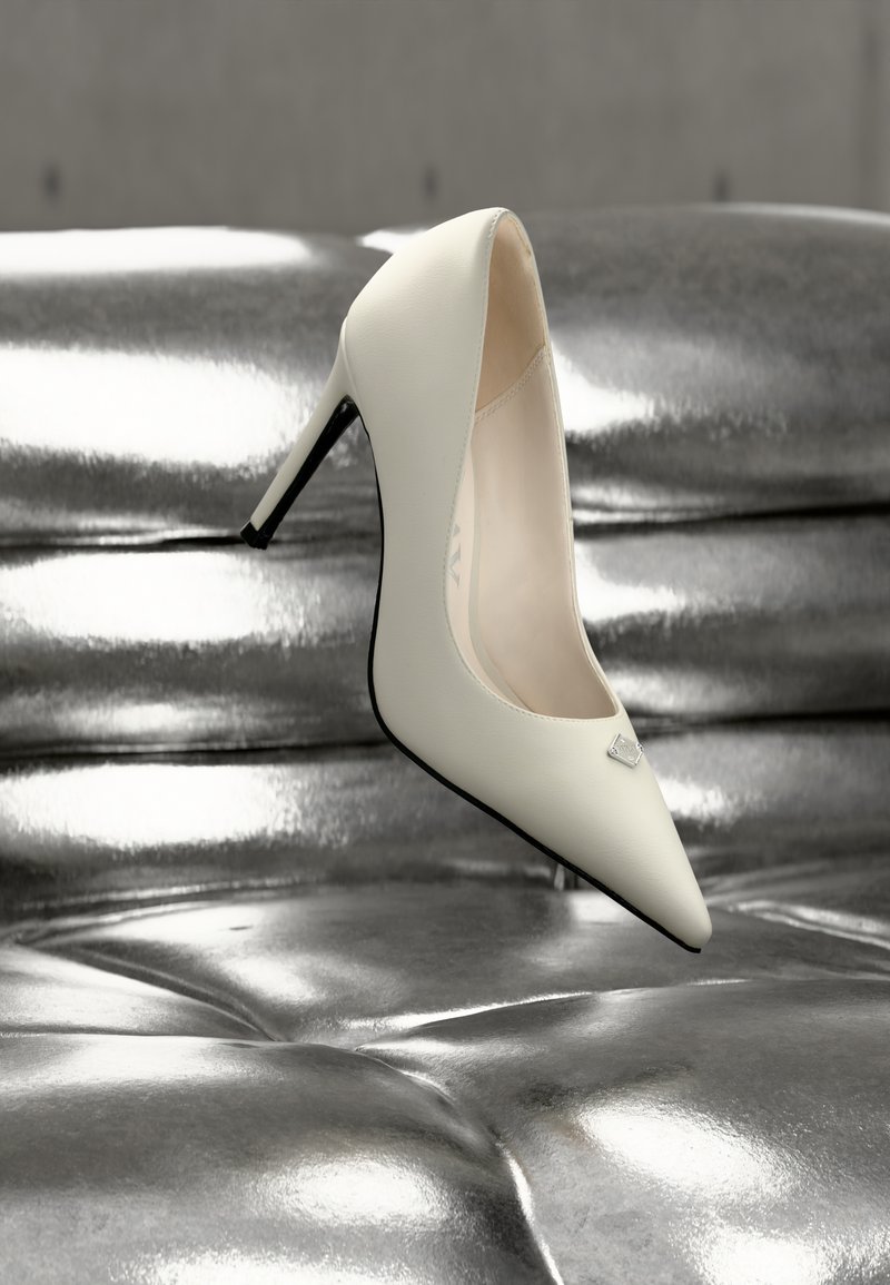 Άσπρο γοβάκι με μυτερή μύτη και κομψό σχεδιασμό, κατασκευασμένο από λείο δέρμα, με μαύρη λεπτομέρεια στη φτέρνα και ένα μικρό ασημένιο έμβλημα.