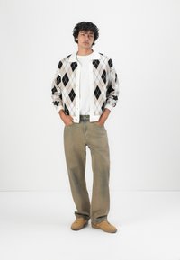 Men's argyle cardigan in cream, black, and beige, paired with a white t-shirt and light khaki loose jeans; smooth texture and relaxed fit.