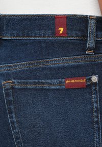 Jean en denim bleu foncé avec des coutures jaunes. Dispose d'une étiquette bordeaux avec un "7" doré et le texte « for all mankind ». Design classique des poches.