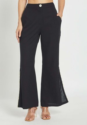 Pantalones negros de pierna ancha con detalles de botones en el lateral, usados con tacones de tiras transparentes, mostrando desde la cintura hasta los pies de una persona de pie con la mano en el bolsillo.