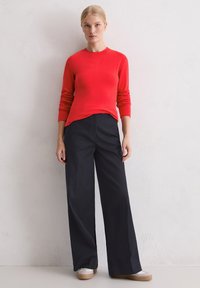 Czerwony sweter z długim rękawem w połączeniu z czarnymi, szerokimi spodniami z wysokim stanem oraz beżowymi butami wsuwanymi. Gładka tekstura; casualowy design.