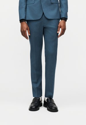 Schmal geschnittene blaue Anzughose, kombiniert mit schwarzen Lederloafers und einer passenden blauen Anzugjacke an einer stehenden Person.