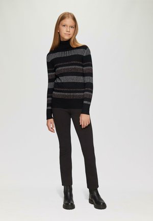 Pullover à col roulé noir à rayures avec texture côtelée et accents métalliques, associé à un pantalon slim noir et des bottes noires épaisses.