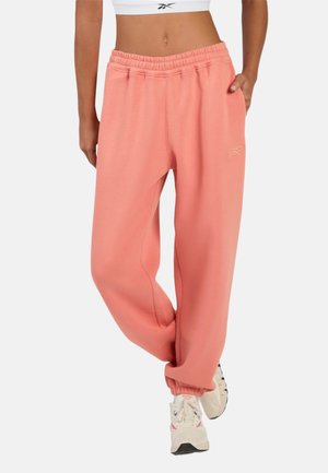 Model draagt koraalroze Reebok joggingbroek met elastische taille en boorden, gecombineerd met witte sneakers en een cropped wit topje.