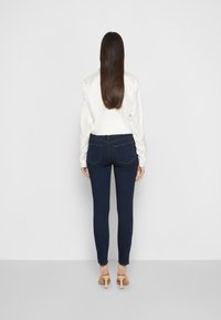 Vaqueros ajustados en azul oscuro, con bolsillos traseros y costuras claras, combinados con una blusa de satén crema y tacones beige.