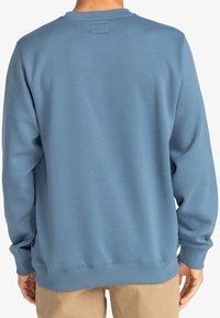 Sudadera azul claro con cuello redondo, puños y dobladillo acanalados, textura suave y una pequeña etiqueta de tela en la parte inferior izquierda.