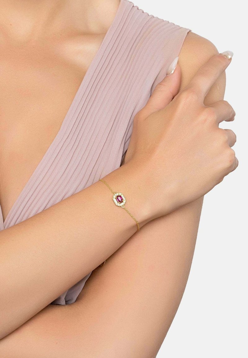 Bracciale d'oro con un centro in pietra preziosa rossa, caratterizzato da un design ottagonale e una delicata catena, indossato su un braccio con pelle chiara.