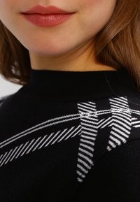 Czarny sweter z wysokim kołnierzem i charakterystycznym białym paskowanym wzorem na ramionach. Miękka tekstura i dopasowany krój.