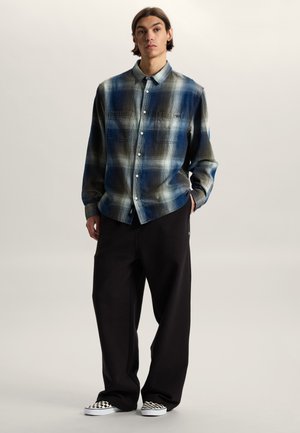 Jeune homme debout, les mains dans les poches, portant une chemise à boutons à carreaux bleus et gris, un pantalon noir ample et des chaussures slip-on à carreaux noir et blanc.