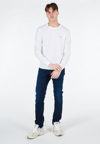 Camicia bianca a maniche lunghe con piccolo logo, abbinata a jeans blu. Le scarpe sono sneakers chiare con dettagli texturizzati.