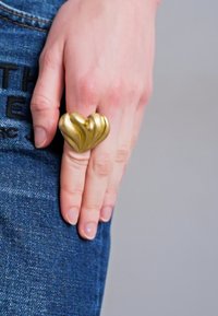 Anello a forma di cuore tono oro con superficie liscia e strutturata, indossato su un dito su uno sfondo di denim.
