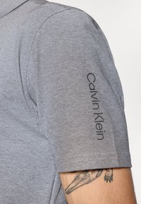 Camisa cinza de mangas curtas com um padrão texturizado. Logo "Calvin Klein" impresso em preto na manga direita. Design e ajuste casuais.