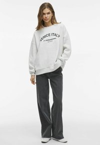 Svetlo siva dukserica s grafiko "VENICE ITALY" v črni barvi, sproščen kroj, dolgi rokavi, v kombinaciji s temnimi širokimi jeans hlačami in belimi čevlji.