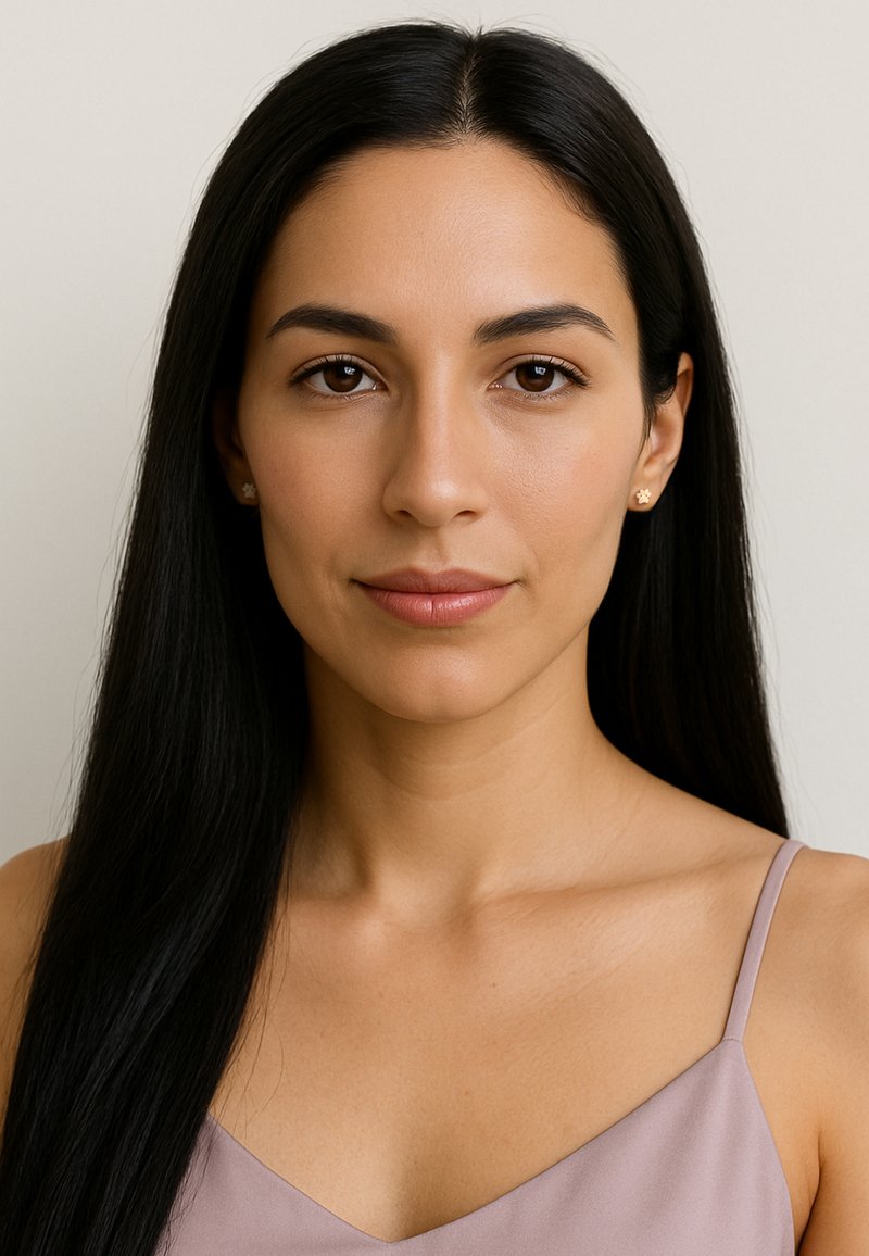 En kvinna med långt svart hår, klädd i en ljuslila satinblus, står mot en enfärgad bakgrund och visar upp minimal smyckespynt och naturlig makeup.