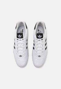 Λευκά αθλητικά παπούτσια με μαύρες ρίγες, με λείο δερμάτινο επάνω μέρος, στρογγυλή μύτη και κλασικό λογότυπο στη γλώσσα και στο πλάι.