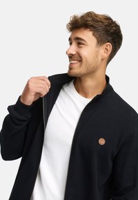 Joven sonriendo, vistiendo una camiseta blanca y desabrochándose una chaqueta de canalé oscura con un pequeño parche de cuero circular.