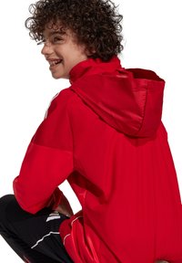 Rote Windjacke mit Kapuze, mit einer glatten Textur und kontrastierenden schwarzen Hosen. Das Design umfasst weiße Akzente an den Ärmeln.