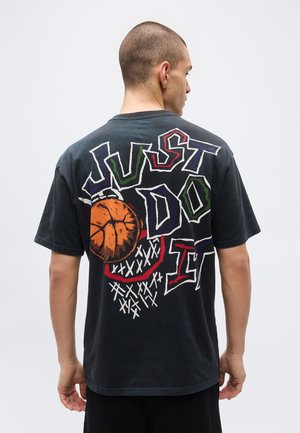 Homme vu de dos portant un t-shirt noir avec le texte coloré « JUST DO IT » et un motif de basketball près d’un panier dans le dos.