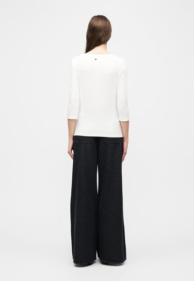 Γυναίκα με μακριά καστανά μαλλιά στέκεται με την πλάτη, φοράει λευκή μπλούζα με μανίκια τριών τετάρτων και μαύρο παντελόνι με φαρδιά μπατζάκια.