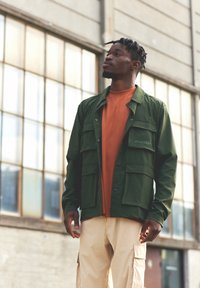 Zelená lehká bunda s několika kapsami přes oranžovou košili, doplněná béžovými cargo kalhotami. Pozadí tvoří industriální okna.